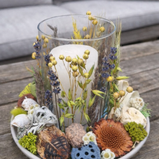 Prírodný sušený aranžmán so sviečkou v skle v zeleno-modrých a hnedých tónoch, doplnený sušenými kvetmi a prírodnými plodmi, položený na drevenom stole.