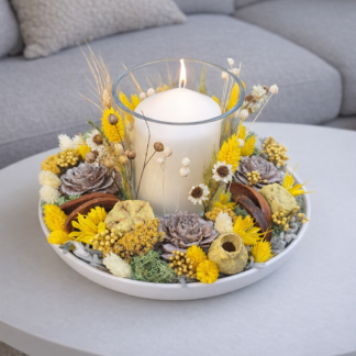Moderný prírodný svietnik s bielou sviečkou v sklenenom pohári, aranžovaný v plytkej miske so sušenými žltými kvetmi, šiškami a prírodnými doplnkami na sivom stolíku pred gaučom.