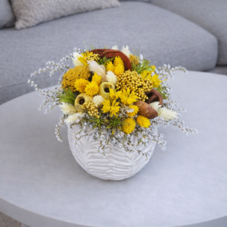 Prírodný aranžmán zo sušených žltých a bielych kvetov v bielom keramickom kvetináči, doplnený sušenými plodmi a prírodnými dekoráciami na sivom konferenčnom stolíku.