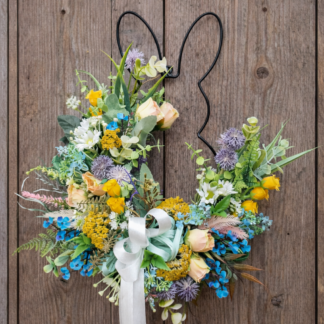 Jarný veniec na dvere v tvare zajačika s farebnými kvetmi a stuhou, zavesený na drevených dverách.